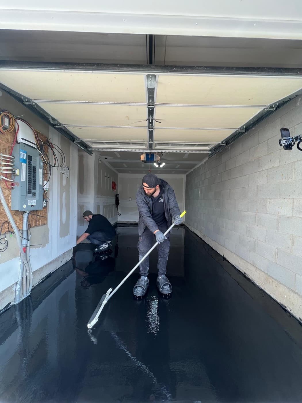 Two LiquidLux installers spreading epoxy across a garage floor with squeegees, garage door open to the street